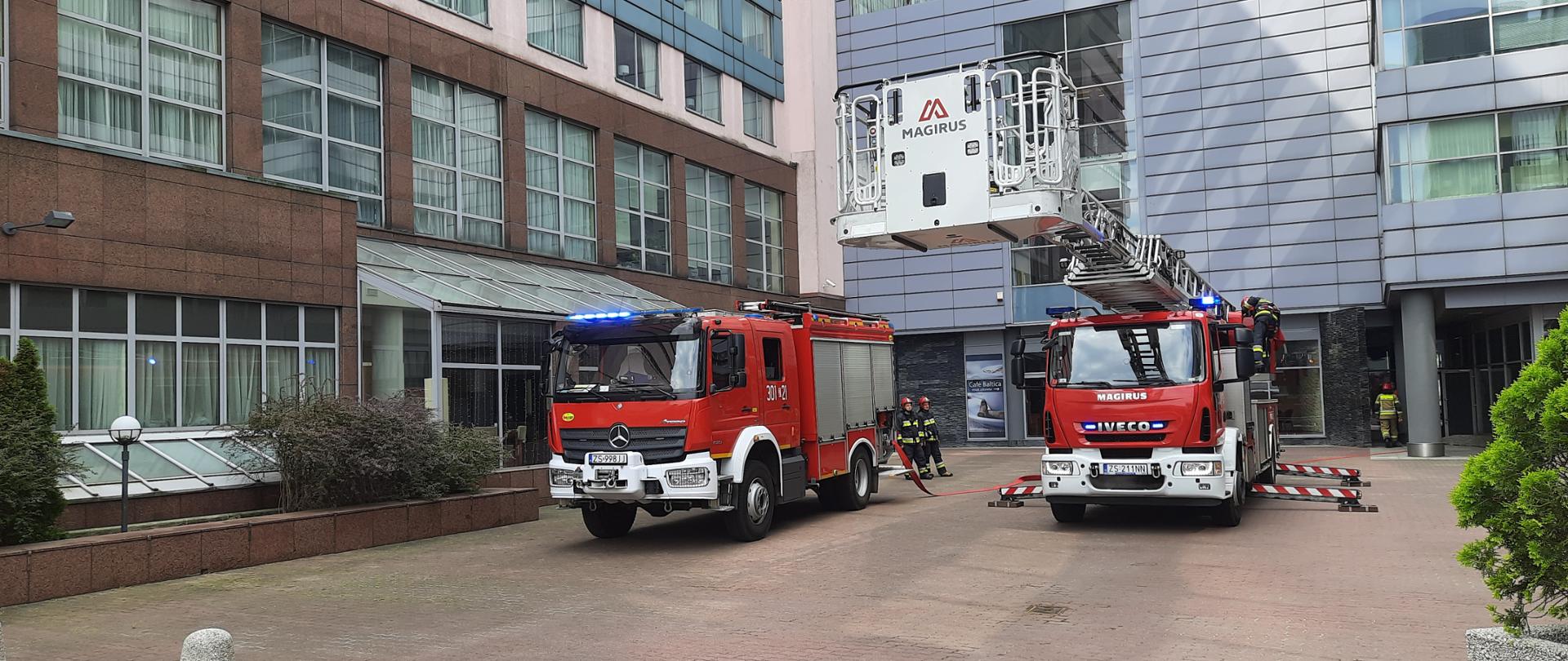 Dwa wozy staży pożarnej w tym jeden to wóz specjalny z drabiną mechaniczną przed kompleksem biurowo hotelowym. Pomiędzy samochodami widać dwóch strażaków w ubraniach specjalnych. 