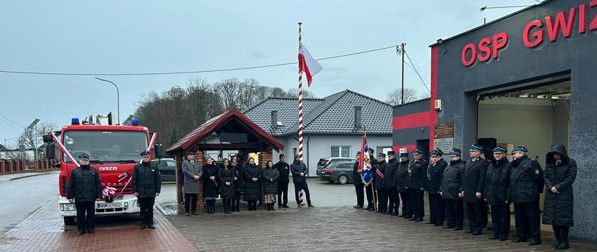 Widok na plac, na którym odbywała się uroczystość. Na zdjęciu samochód pożarniczy, uczestnicy uroczystości i pododdział druhów OSP Gwiździny.