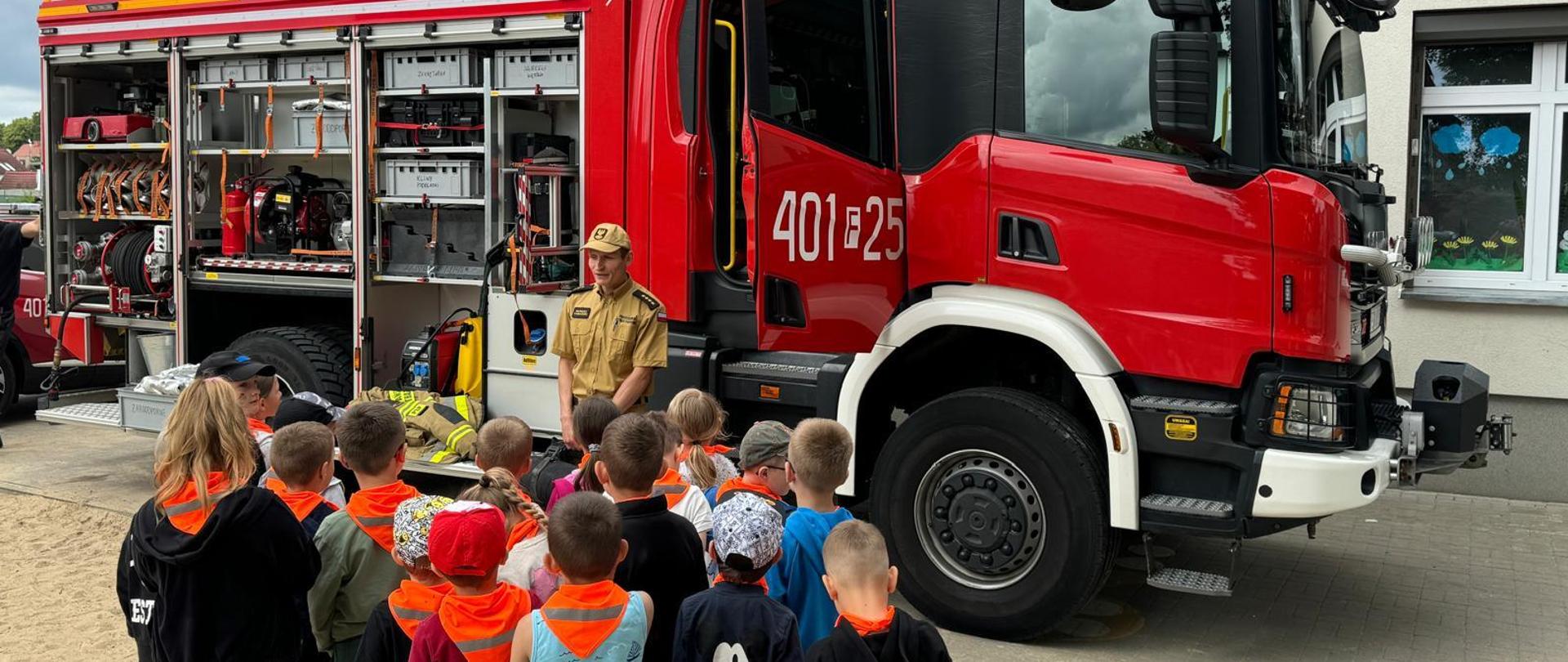 Zdjęcie przedstawia grupę przedszkolaków słuchającą strażaka. Na drugim planie znajduje się samochód pożarniczy.