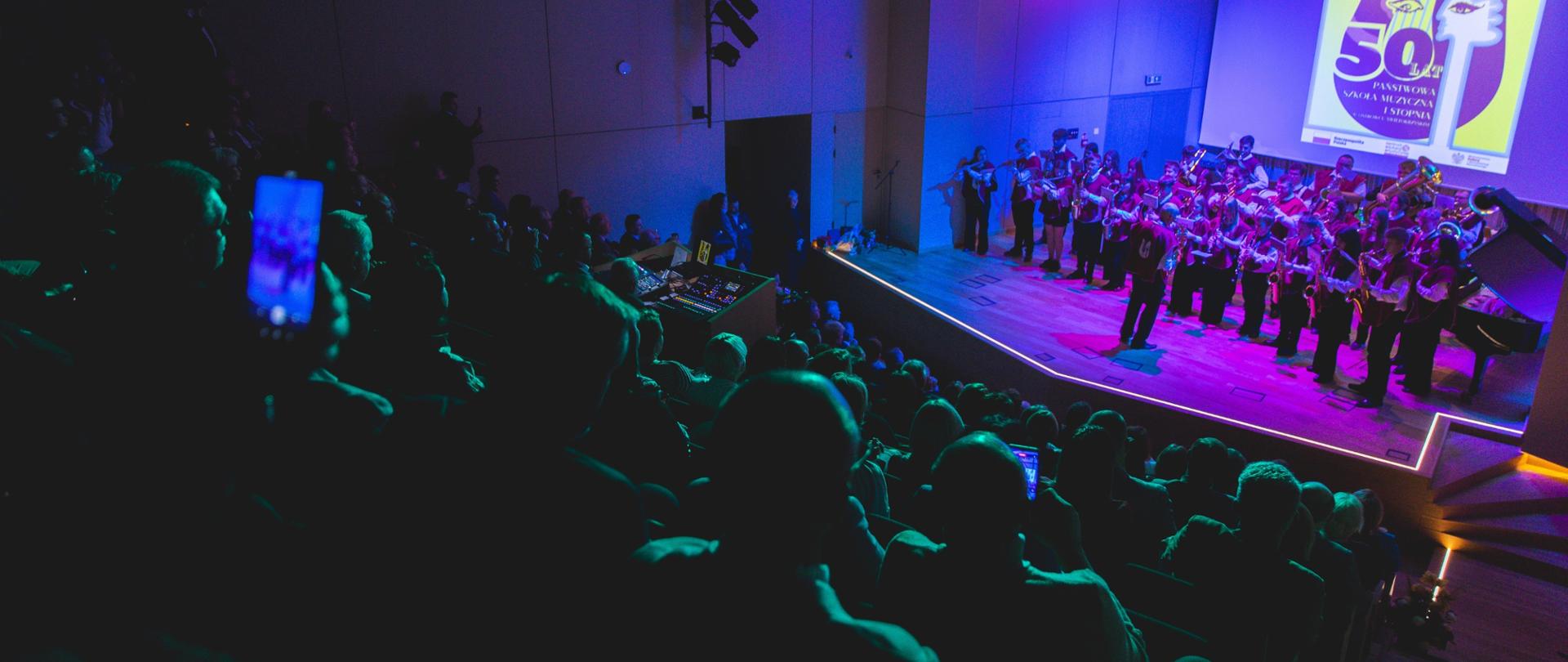 koncert orkiestry na scenie, obserwująca publiczność, w tle logo szkoły 