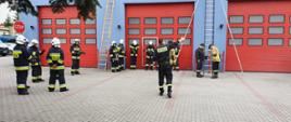 Zdjęcie przedstawia strażaków w umundurowaniu specjalnym na placu komendy powiatowej PSP w Jędrzejowie. O budynek garażowy jednostki ratowniczo-gaśniczej oparte są dwie drabiny. Strażacy przygotowują się wspinania się po drabinach.
