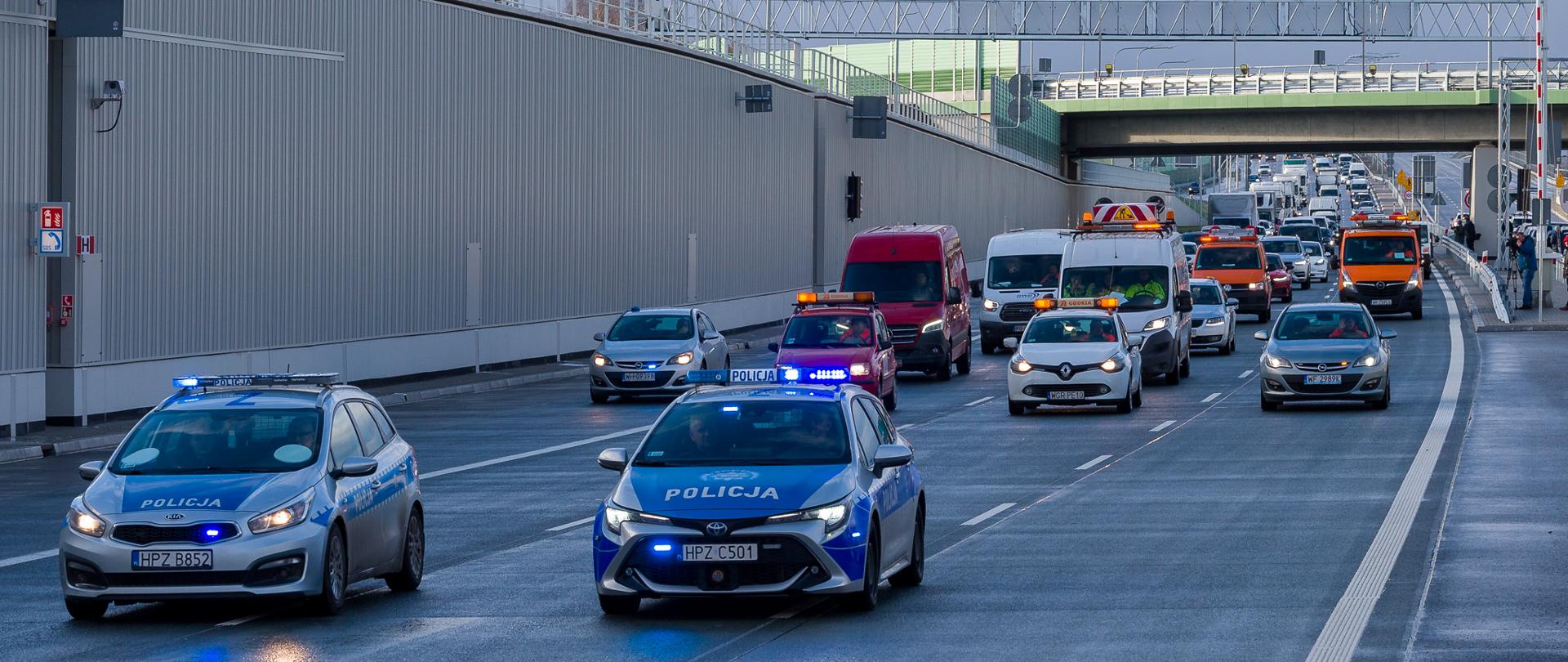 Zdjęcie przedstawia scenę na wielopasmowej drodze z wieloma pojazdami, w tym dwoma radiowozami z migającymi niebieskimi światłami, czerwonym vanem oraz kilkoma innymi samochodami i pojazdami serwisowymi GDDKiA. Wszystkie pojazdy poruszają się za dwoma radiowozami na pierwszym planie.